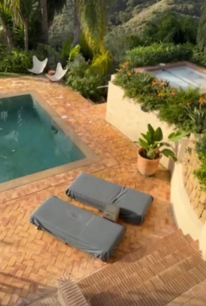 Zona de piscina con tumbonas grises y plantas, rodeada de vegetación.