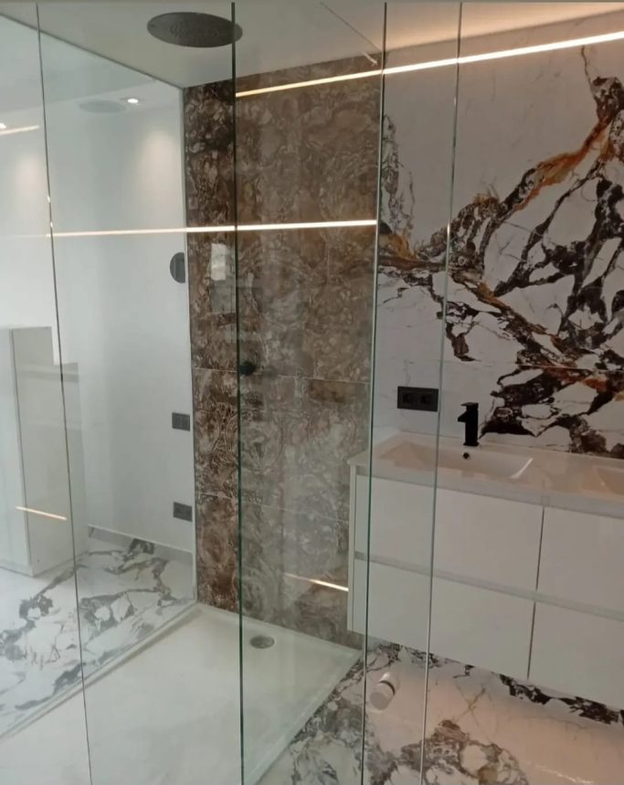 Baño moderno con ducha de cristal, paredes de mármol y lavabo blanco.