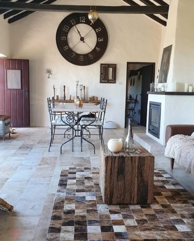 Sala de estar acogedora con una mesa, sillas, reloj grande y una alfombra de patchwork.