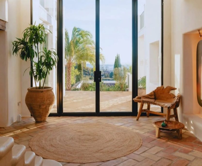 Entrada luminosa con plantas, suelo de barro y grandes ventanales hacia el jardín.
